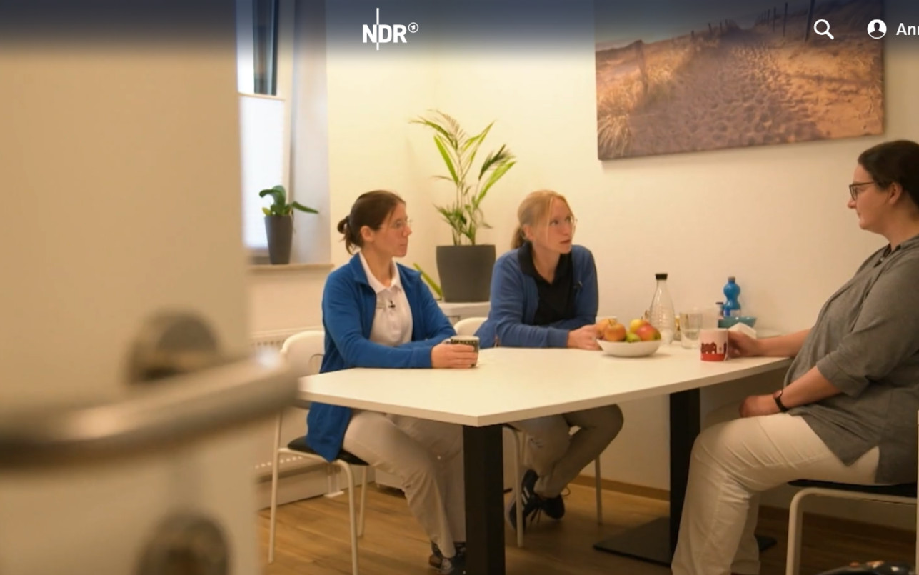 Screenshot aus dem Video: Blick durch eine Tür auf unsere Ärztinnen, die sich am Tisch sitzend unterhalten.